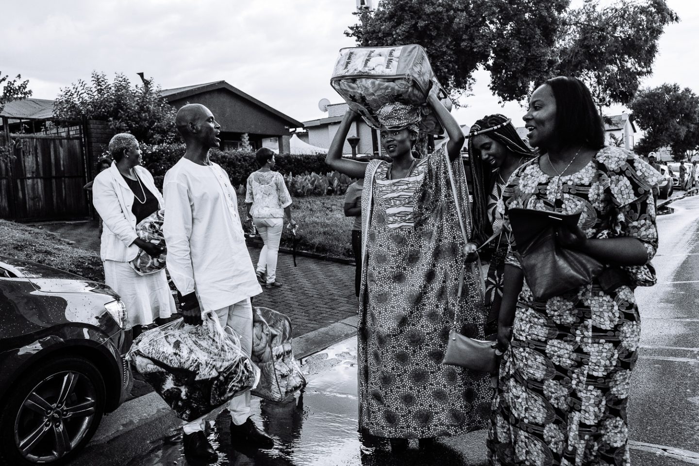 Guests depart carrying gifts on a wet street outside a family home. Johannesburg, South Africa, 2017.