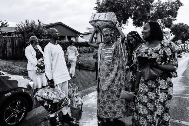 Guests depart carrying gifts on a wet street outside a family home. Johannesburg, South Africa, 2017.